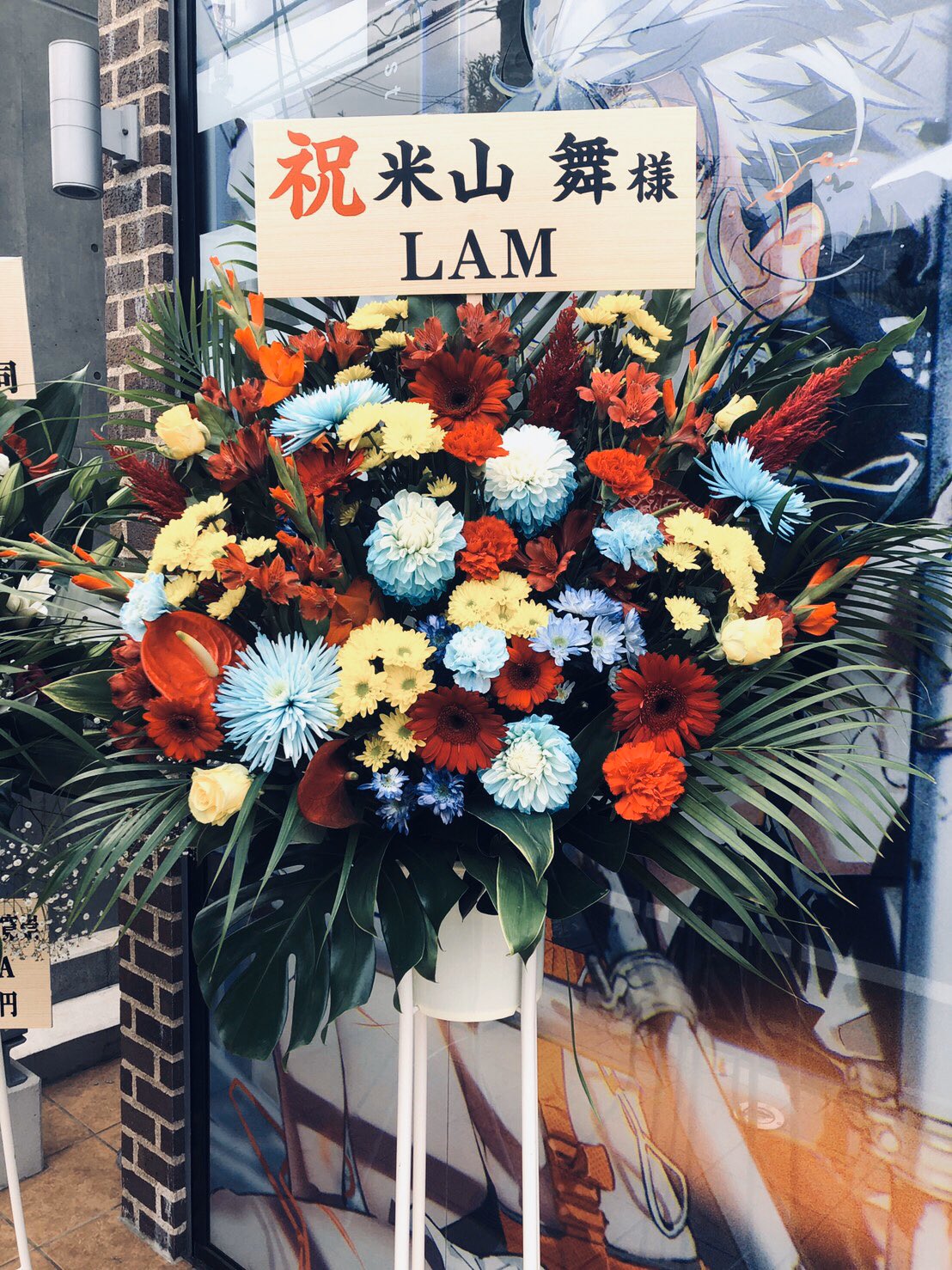 a large floral arrangement, predominantly in red, blue, yellow, and white, stands on a sidewalk in front of a storefront.  the arrangement, a traditional japanese celebratory gift, likely commemorates a store opening or other special occasion.  the sign on the arrangement features japanese writing and the roman letters "lam."