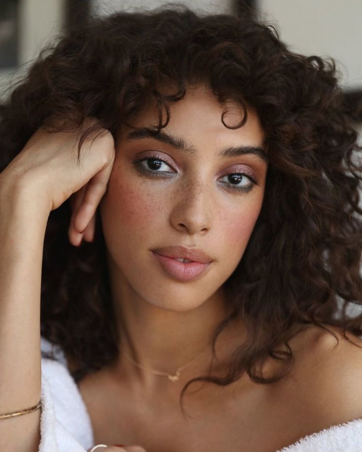 the image shows a closeup portrait of a woman with curly brown hair and freckles. she has light pink blush and eyeshadow, and wears a delicate gold necklace and bracelet. she is resting her head on her hand, and appears relaxed. she is wearing a white towel around her shoulders.
