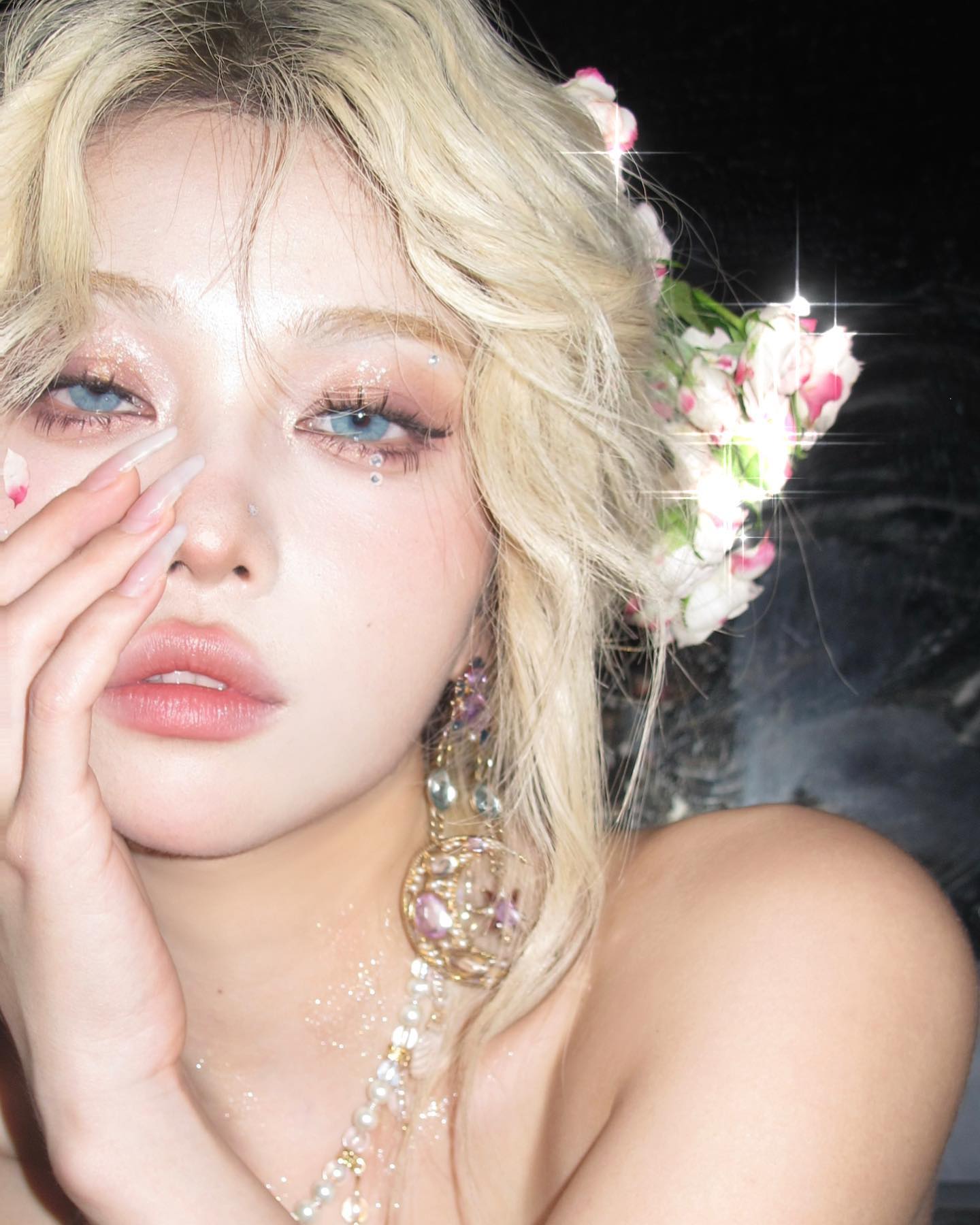 a young woman with blonde hair and blue eyes poses for a closeup selfie. she has a flower crown in her hair and is wearing delicate jewelry. her makeup is ethereal and she has a captivating gaze. the lighting is soft and the overall aesthetic is dreamy and feminine.