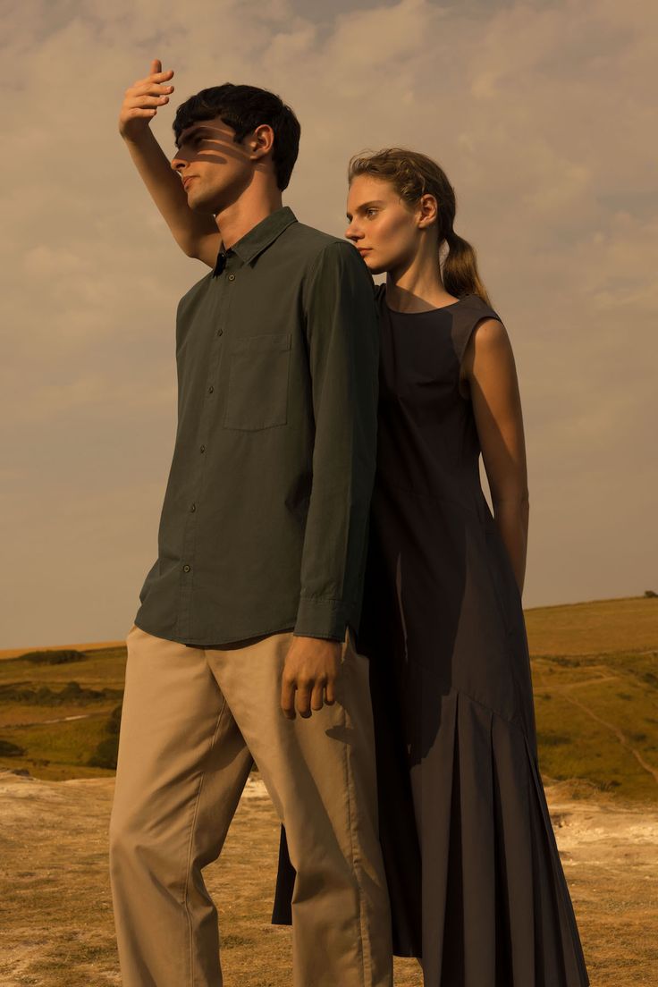a man and a woman stand in a field. the man is wearing a longsleeved green shirt and brown pants. he is shielding his eyes from the sun with his hand. the woman is wearing a sleeveless blue dress. they are both looking at the camera.