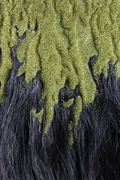 a closeup shot of a surface of long black strands of hair interwoven with green strands that are arranged in an abstract and repeating pattern.