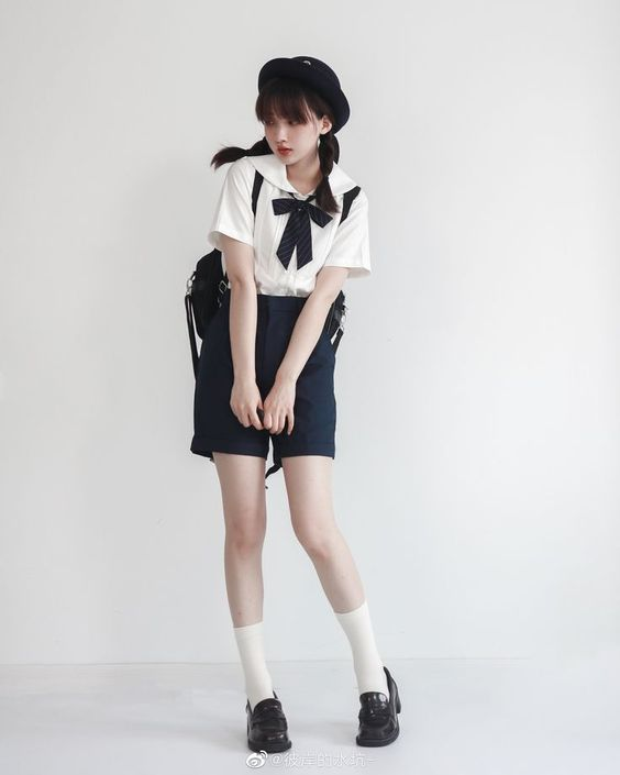 a young woman poses against a white background, modeling a stylish outfit. she wears a white collared top with a black bow, paired with navy blue shorts. her accessories include a black beret, a black crossbody bag, white socks, and black loafers. the image captures a clean and fashionable aesthetic with soft lighting.