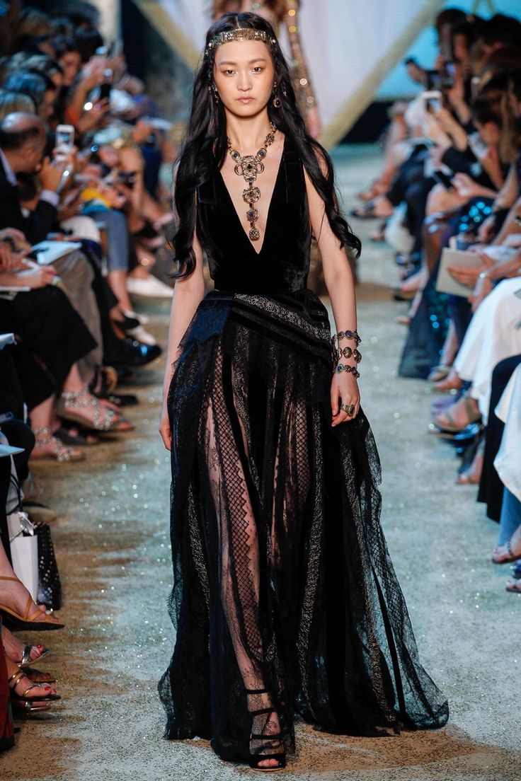 the image showcases a highfashion runway model walking down a catwalk. she is wearing a stunning black floorlength gown, which is designed with a velvet bodice and a sheer lace overlay. the dress includes intricate details. the model also sports several ornate pieces of jewelry, including a large statement necklace, a headband, bracelets, and rings. her long, dark hair flows down her back, complementing the dark and luxurious aesthetic of the overall look. the background is blurred, showing a seated audience at a fashion show, further emphasizing the model and her attire. the overall ambiance is one of elegance, sophistication, and high fashion.