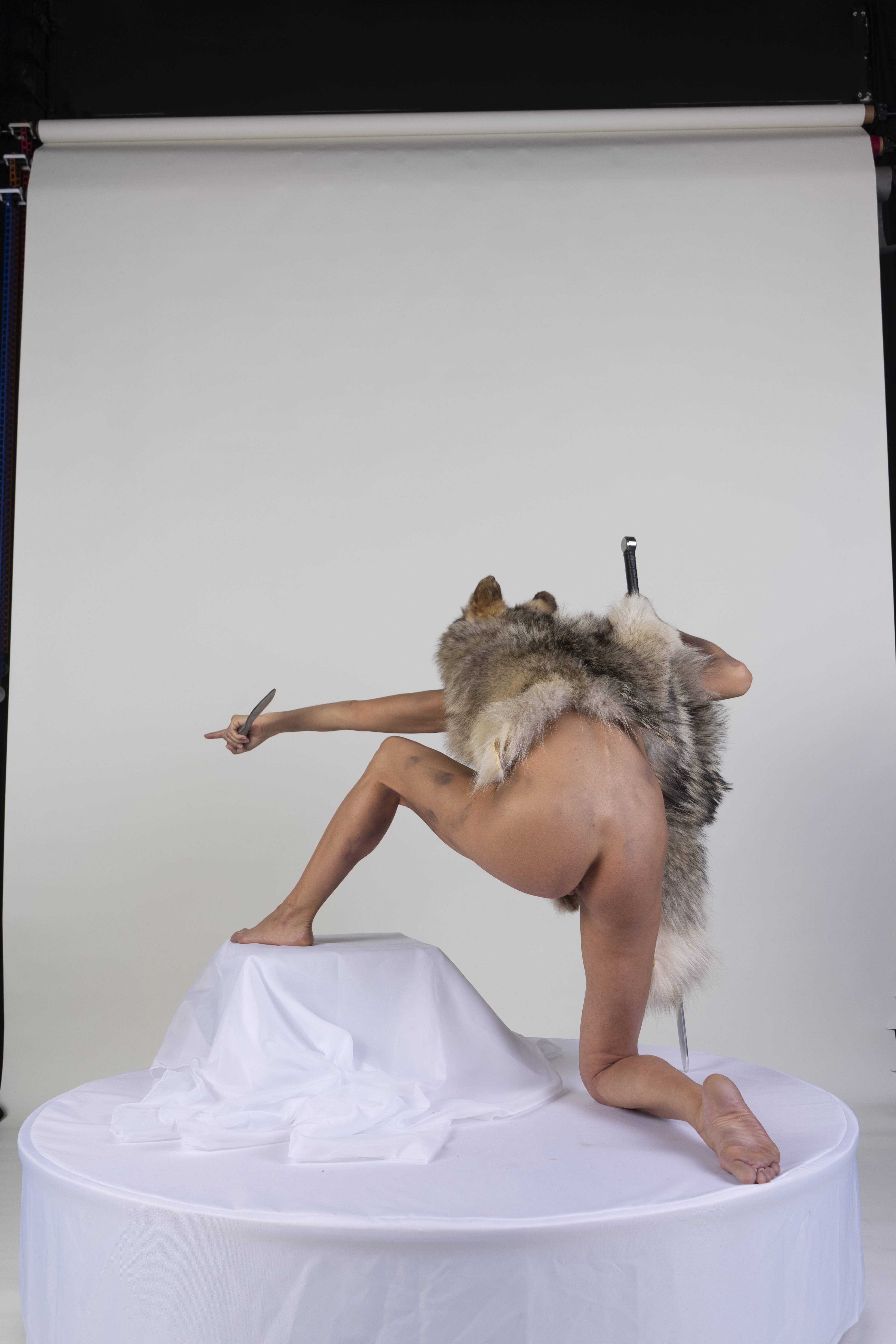 a nude woman wearing a wolf pelt and holding a knife is kneeling on a white platform in a studio with a white background. she has a sword on her back.