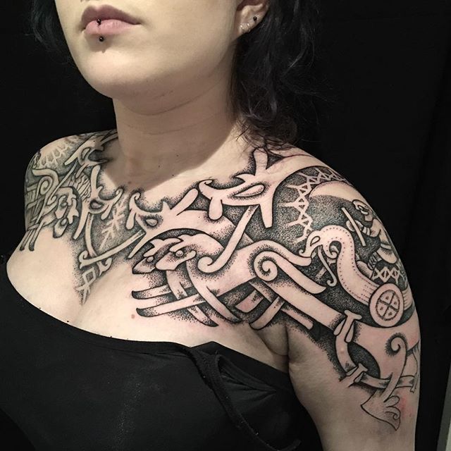 a woman with a vikingthemed tattoo on her upper chest and shoulder. the tattoo features intricate knotwork, animals, a viking figure in a boat, and possibly an axe. the tattoo style is primarily black ink with dotwork shading. the woman has lip and ear piercings. she is wearing a black top.
