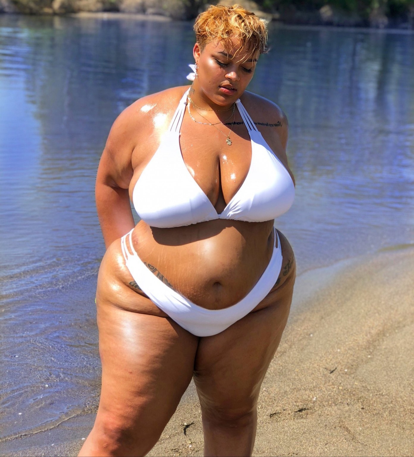 a beautiful woman in a white bikini stands on a sandy beach with the ocean behind her. she is looking down and smiling, and she looks very happy and relaxed. the sun is shining and the water is sparkling. the woman is wearing a white bikini top with thin straps and a white bikini bottom with a thin strap along the side.  she is tan, and her body is toned. the woman is in a beautiful place, and she looks like she is having a great time.