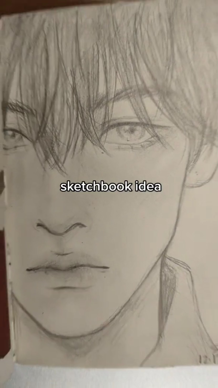 the image shows a pencil sketch of a young man's face. the drawing is done in a realistic style, with careful attention to shading and detail. the subject is looking directly at the viewer with a neutral expression. the words "sketchbook idea" are written across the center of the image.