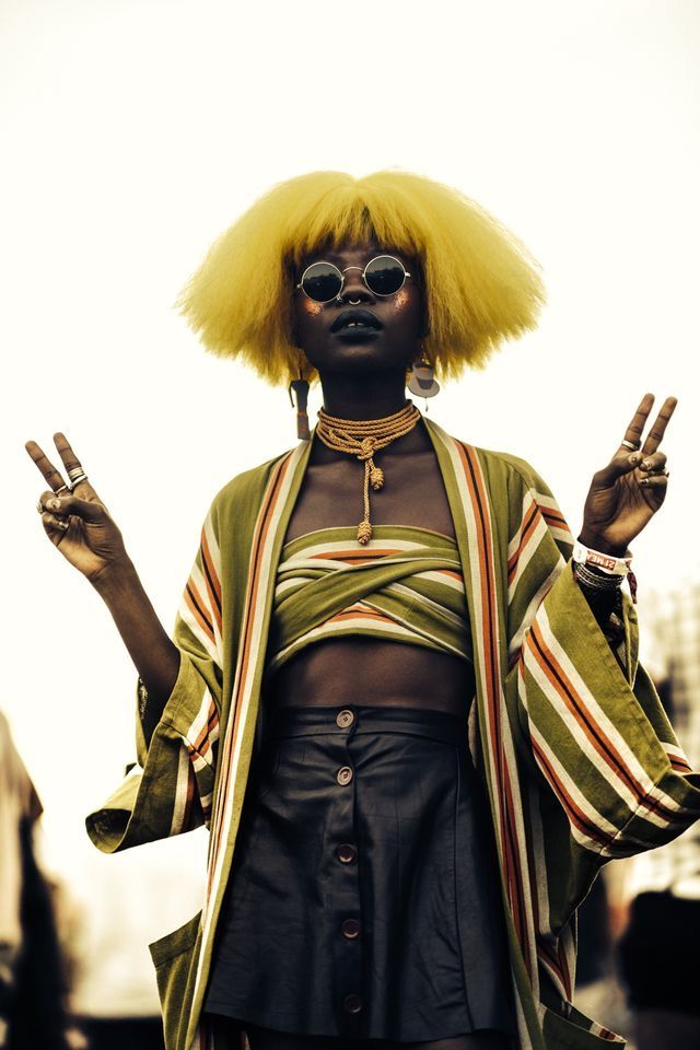 a woman with bright yellow hair and sunglasses is wearing a striped jacket, a crop top, and a black mini skirt. she's making the peace sign with both of her hands.