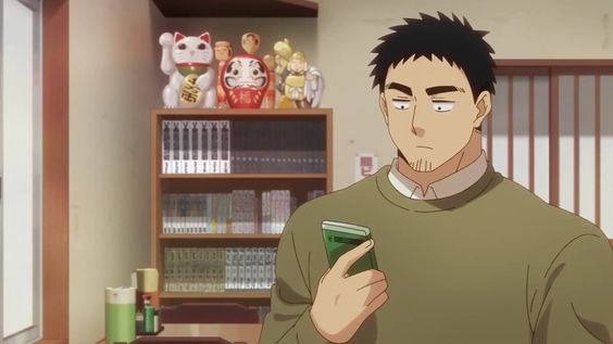 a man with short black hair and a green sweater is looking down at a green phone in his right hand. he is standing inside in front of a bookshelf with various collectibles on it. the lighting is bright and the perspective is at eye level.