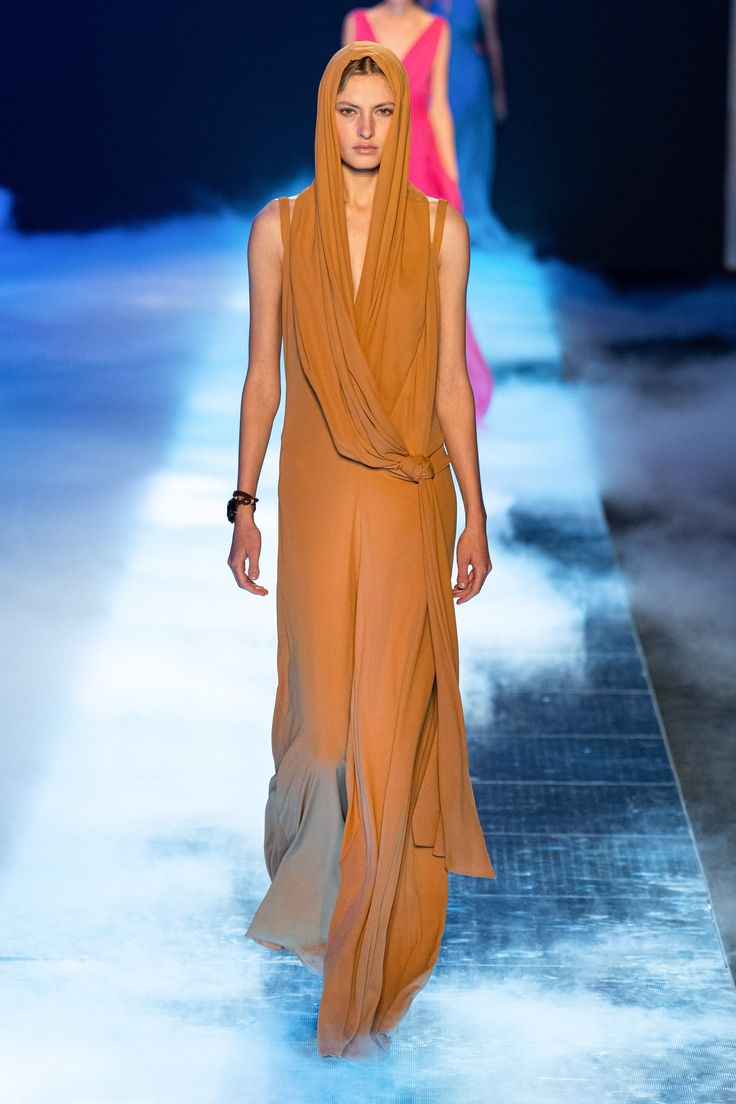 a fashion model walks the runway in a long, flowing brown dress with a cowl neck and a hood. the dress is draped in a way that creates a very elegant and chic silhouette. the model is wearing minimal makeup and her hair is pulled back from her face, allowing the dress to be the focus. the runway is bathed in a soft, diffused light, which creates a sense of drama and mystique. the model is confidently striding towards the camera, her eyes fixed on the horizon.
