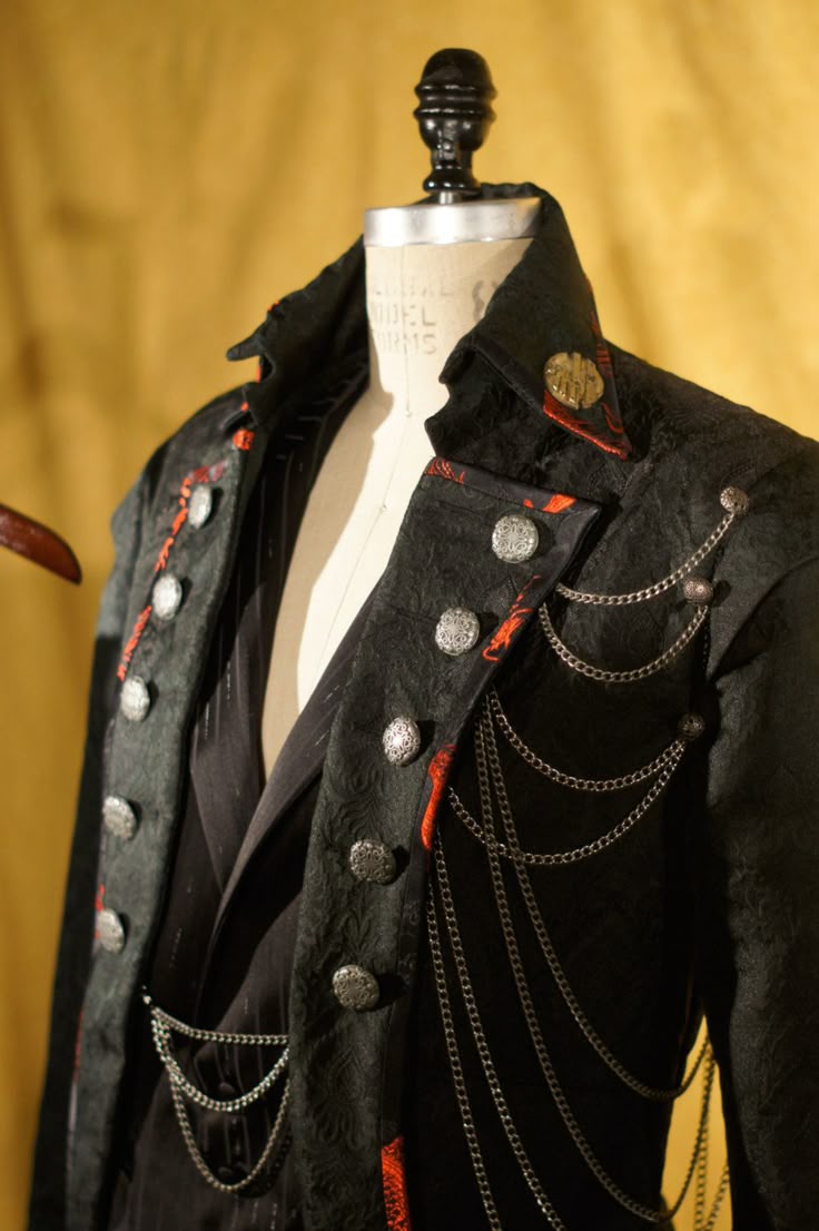 a closeup studio shot showcases a black coat with ornate silver buttons and multiple decorative silver chains cascading down the side. the coat is displayed on a mannequin, and it features a high collar, and red accents. the texture of the coat is detailed and patterned.