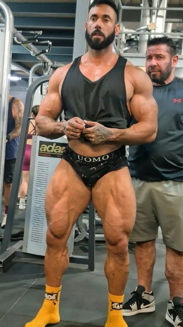 a powerfully built bodybuilder stands in a gym, flexing his legs and showcasing his impressive musculature. he's wearing black underwear with a patterned waistband, and yellow socks with branding.  another man, seemingly a friend or trainer, stands behind him, observing.  the background features gym equipment and partial views of other individuals. the overall ambiance suggests a setting of intense physical training.