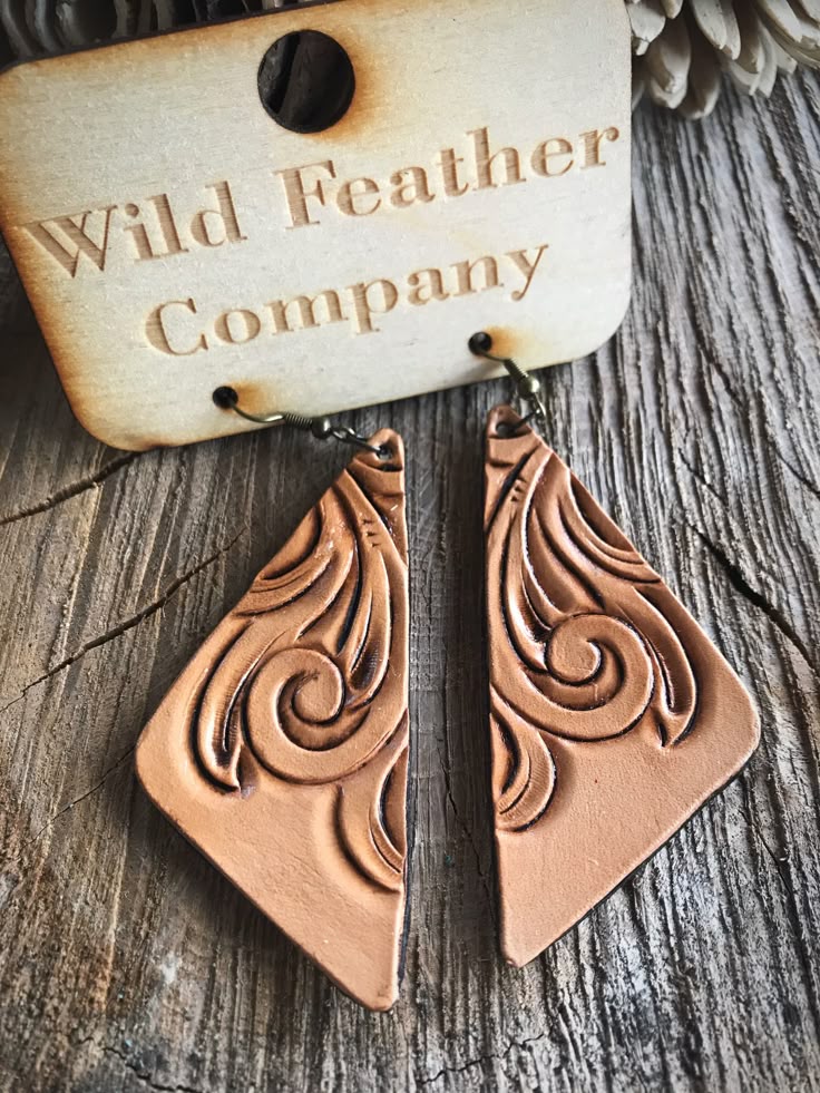 closeup shot of a pair of tan leather earrings with an etched swirl design, hanging from a wooden tag that reads "wild feather company". the earrings are triangular in shape and are placed on a weathered wooden surface.