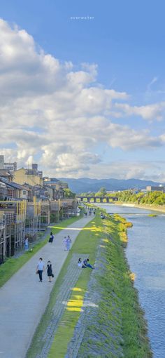 the image captures a picturesque view of a river flowing through a town, likely in japan. traditional wooden buildings line the riverbank, with a paved path running alongside. people can be seen strolling along the path, enjoying the sunny day. a bridge arches over the river in the distance, leading to a range of mountains under a bright blue sky dotted with fluffy white clouds. the overall atmosphere is serene and inviting, showcasing the beauty of japanese architecture and nature in harmony.