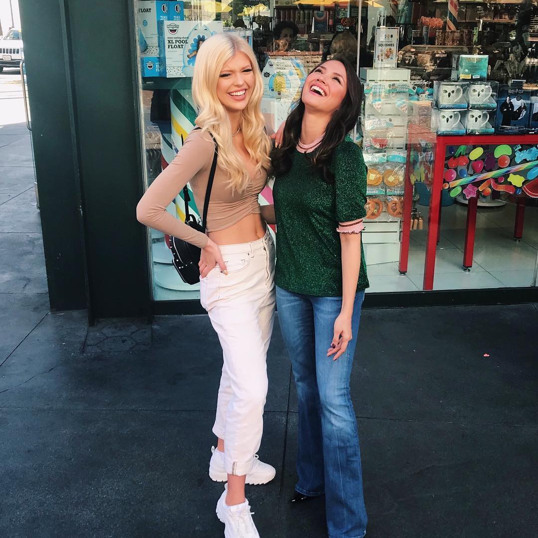 two women are standing outside a shop window, laughing. the woman on the left has blonde hair and is wearing a beige longsleeved shirt, white pants, and white sneakers. she has a black bag on her shoulder. the woman on the right has brown hair and is wearing a green shirt and jeans. they both appear to be happy and enjoying each other's company. the shop window behind them has various items on display.