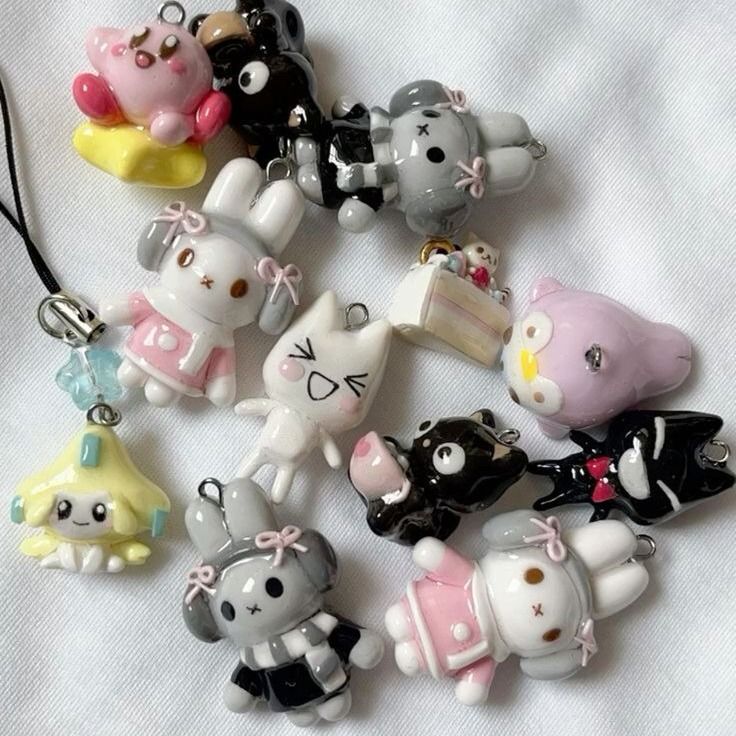 the image shows a collection of handmade polymer clay charms and keychains. there are various character figurines including pink and gray bunnies with bows, black and gray bears, a white cat, a pink character lying down, a yellow starshaped character, a cake with a bear on top, and other abstract shapes in pink and black. the charms are arranged on a white surface.
