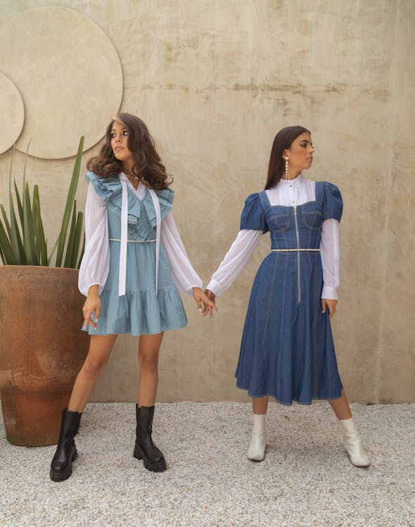 two young women are posing outdoors, holding hands, and wearing denim dresses over white shirts with boots. the woman on the left is wearing a light blue denim dress with ruffles and black boots. the woman on the right is wearing a darker denim dress with puff sleeves and white boots. they are standing on a gravel surface in front of a textured wall with circular design elements. a large plant is visible on the left side of the image.