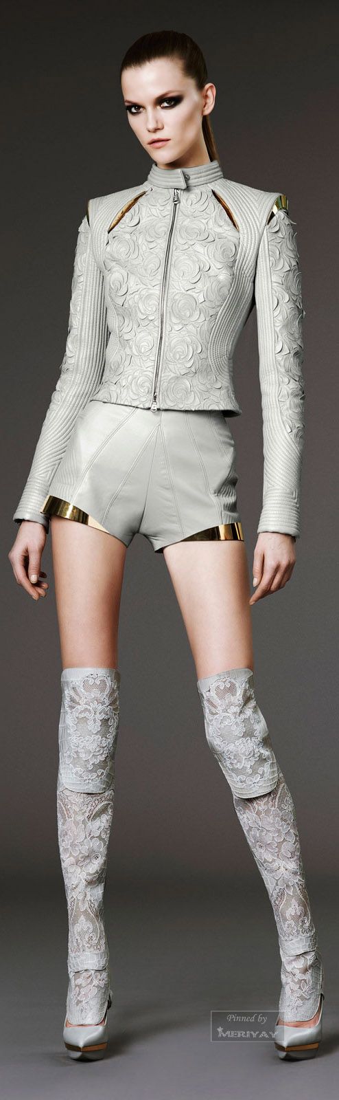 a woman with a serious expression poses in a studio. she is wearing a white leather jacket and shorts, and lace boots. her legs are spread wide, and she's looking straight ahead at the camera. the background is a light grey.