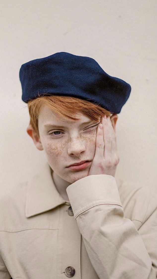 a closeup portrait of a young boy with red hair and freckles wearing a beige jacket and a blue beret. he has his hand on his cheek and is looking down with a serious expression.  the background is a plain white wall.