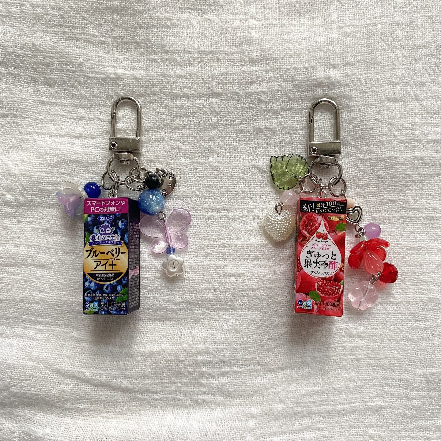 the image shows two keychains on a white textured surface. the keychain on the left features a miniature box of blueberryflavored health supplement, decorated with blue and purple beads, a butterfly charm, and a small heart. the keychain on the right showcases a miniature box of pomegranateflavored fruit vinegar, adorned with a green leaf charm, a pearl heart charm, and pink and red flowershaped beads. both keychains have silvertoned clasps at the top.