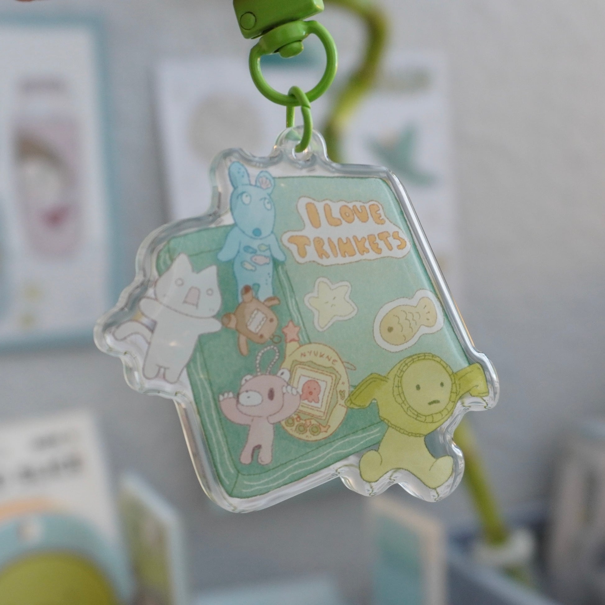 closeup view of a clear acrylic keychain featuring a pastelcolored illustration of various cute cartoon characters and objects arranged around a book that says "i love trinkets."  the characters are soft, rounded shapes, and the overall style is whimsical and kawaii. the keychain is attached to a small green plastic ring, and it appears to be hanging.  the background is blurry, showing other objects and suggesting a home or shop setting.