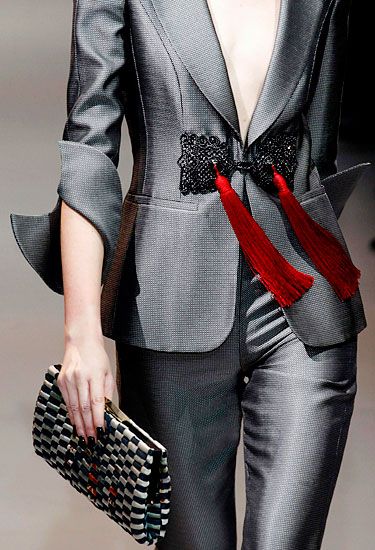 a woman is wearing a grey woman's business suit with a black lace detail on the blazer along with two red tassels. she's carrying a black and white clutch. the woman is facing the camera and the shot is a midshot showing from above her head to her midthigh.