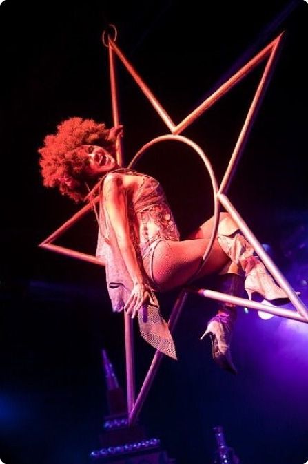 a woman in a pink outfit hangs from a hoop  in a starshaped framework. she's in midair, performing an acrobatic routine. her afro is flowing, and she's smiling. the stage is lit in pink and purple, and the background is dark.