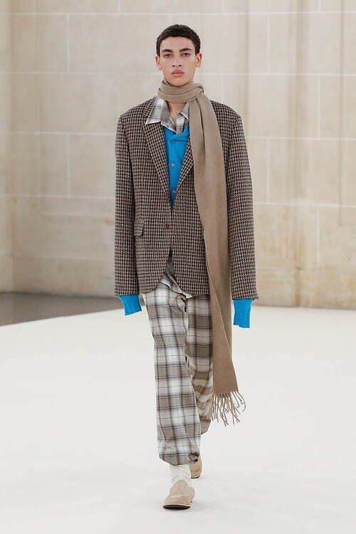 a male model walks the runway during a fashion show wearing a checkered brown and black blazer, plaid pants, a blue sweater, and a tan scarf. he is also wearing white sneakers. the model is walking on a white runway in front of a white wall.