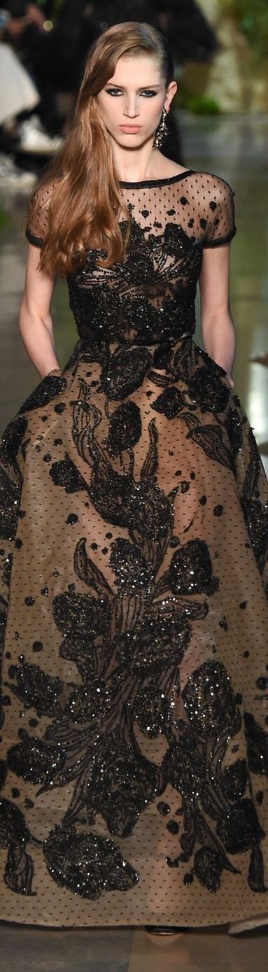a beautiful woman is standing on a runway wearing a long, black, sheer gown with intricate floral embroidery and sequins. the dress is very elegant and glamorous. the model is looking straight at the camera with a slight smile on her face. the dress is very detailed and intricate, with a variety of textures and patterns. the model's hair is styled in a loose, wavy style. the dress is perfect for a formal occasion.