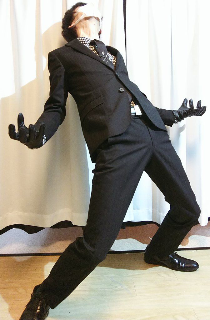 a man in a black suit with a white tie and gloves stands in front of a white backdrop. his body is contorted into a dramatic pose, with one hand extended outward and the other bent at the elbow. he looks up towards the sky with his eyes closed. the man is dressed in a formal outfit, with a black suit jacket and pants and a white tie. his black gloves add a touch of mystery to his look. the overall tone of the image is dark and stylish.