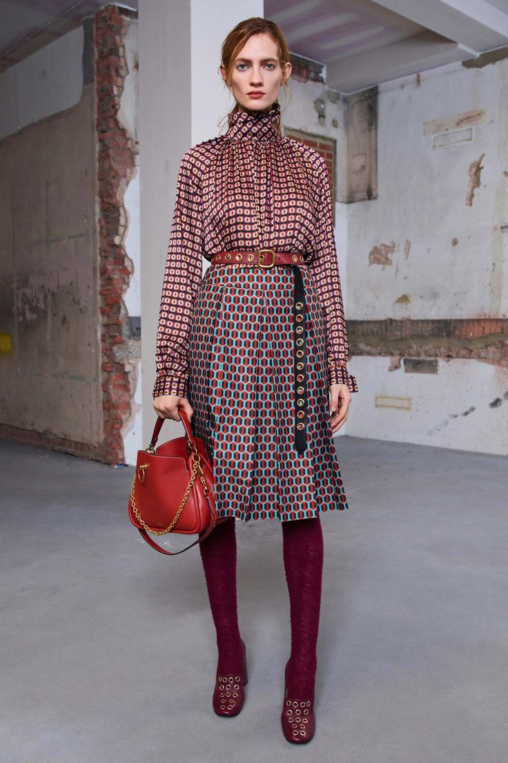 a full shot of a fashion model showcasing an autumnal outfit. the model, a woman with light skin and reddishbrown hair styled in a low bun, is wearing a kneelength pleated skirt and a matching longsleeved blouse with a geometric pattern in shades of burgundy, red, and teal. the outfit is cinched at the waist with a brown belt with gold accents. she also sports burgundy tights and burgundy loafers with decorative eyelets.  the model carries a red leather shoulder bag. the setting is an industrialstyle interior with a distressed brick wall in the background. the overall impression is one of sophisticated, autumnal style.