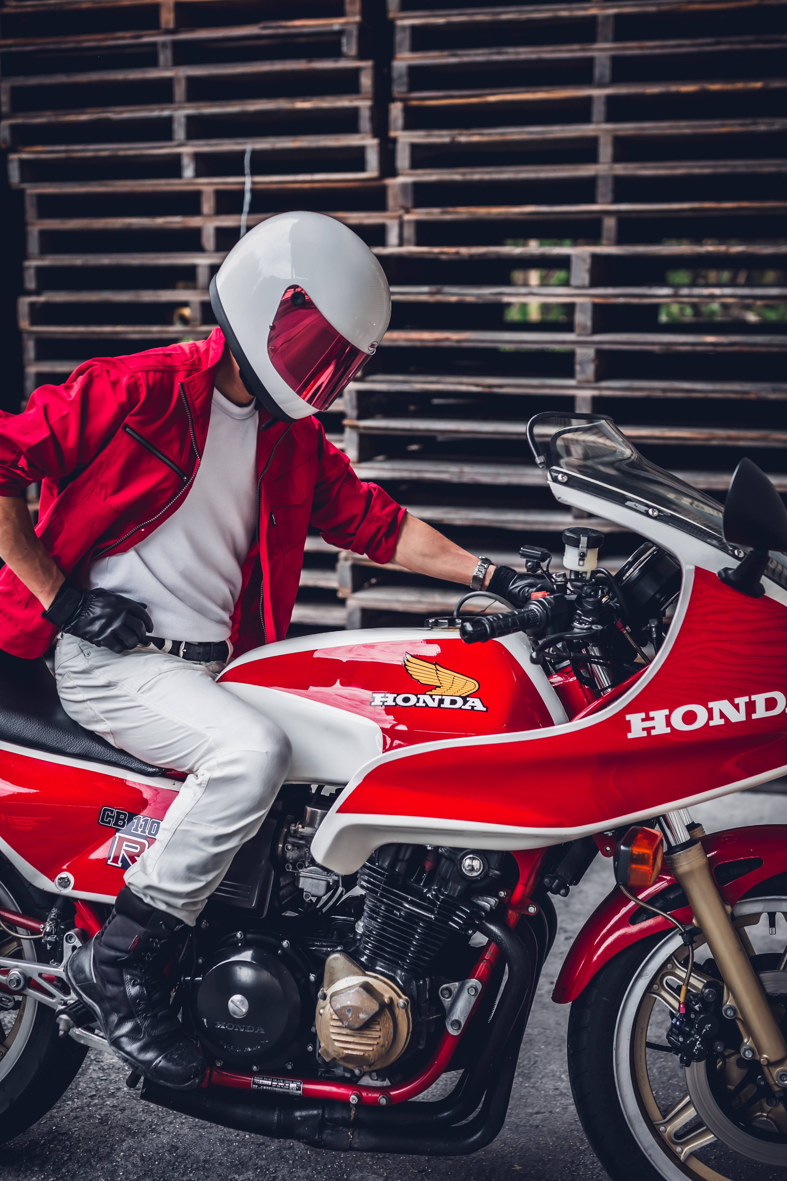 a man wearing a red jacket and white pants sits on a red and white honda motorcycle with a vintage white helmet.  the motorcycle is parked in front of a wooden pallet wall. the man is looking down at the motorcycle. the motorcycle is in a classic style.