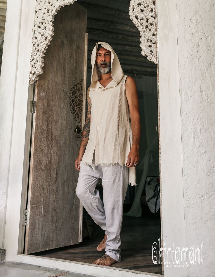 the image shows a man, possibly a model, wearing a beige linen or cotton bohemianstyle outfit. the outfit consists of a sleeveless hooded tunic and loosefitting lightgrey pants. he is barefoot and standing in a doorway with an ornate, carved archway. the setting is minimalist and serene, with white walls and a simple wooden door. the overall vibe is peaceful and spiritual, suggesting a lifestyle brand or a fashion shoot for clothing related to yoga, meditation, or spiritual practices.  a watermark for "chintamani alchemy" is visible in the bottom right corner.