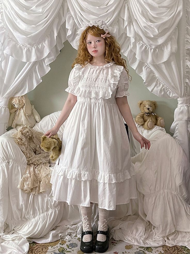 a young woman with long, curly red hair stands in a room filled with white, ruffled fabric. she is wearing a white, vintageinspired dress with delicate lace detailing and ruffles. her hair is adorned with a white bow. two teddy bears rest on a chaise lounge behind her, draped in similarly textured white fabric. the overall ambiance is soft, dreamy, and romantic. the image has a vintage quality, reminiscent of antique dolls and victorian aesthetics.