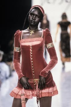a black female model walks down a runway in a red velvet dress with a pink lace trim. she is wearing black gloves and a gold belt. she is posing for the camera.