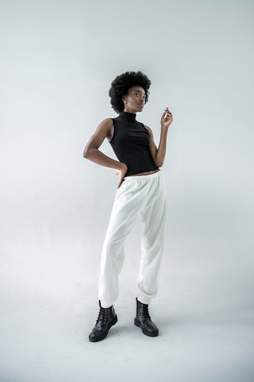 a full studio shot of a slender, tall black woman standing against a white background, looking off to the right with an air of confidence. she has a large afro and is wearing a black, sleeveless turtleneck top, white sweatpants that are slightly baggy, and black boots. her left hand is on her hip, while her right hand is raised with her fingers extended. she's also wearing earrings. the lighting is even, highlighting her features and the texture of her clothes. the overall style is casual and fashionable.