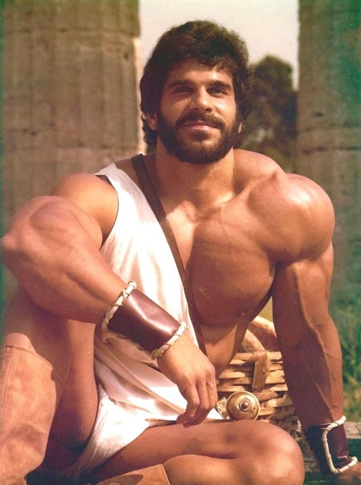 a vintage photograph depicts a heavily muscled, tanned man with a dark beard and curly hair. he's dressed in a simple white togalike garment and leather armbands.  he's seated amidst the ruins of what appear to be ancient greek columns, holding a small basket partially in his lap. his powerful physique is prominently displayed, suggesting a bodybuilder or fitness model. the photograph has a slightly warm and faded quality typical of vintage prints. the overall mood is one of strength, masculinity, and classical aesthetics.