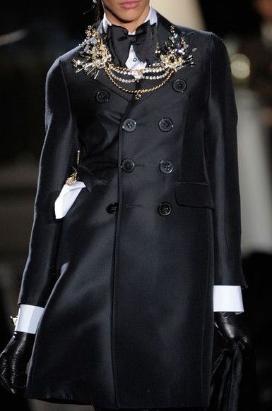 closeup shot of a fashion model showcasing a stylish black doublebreasted coat. the coat features a sleek design with clean lines and is accessorized with a layered pearl and gold necklace, a black bow tie, and black gloves with white cuffs. the model's expression is serious and poised, enhancing the overall sophisticated and elegant feel of the image, suitable for a highfashion magazine or website. the background is blurred, focusing attention on the coat and the model's attire. the image suggests a fall or winter fashion collection.
