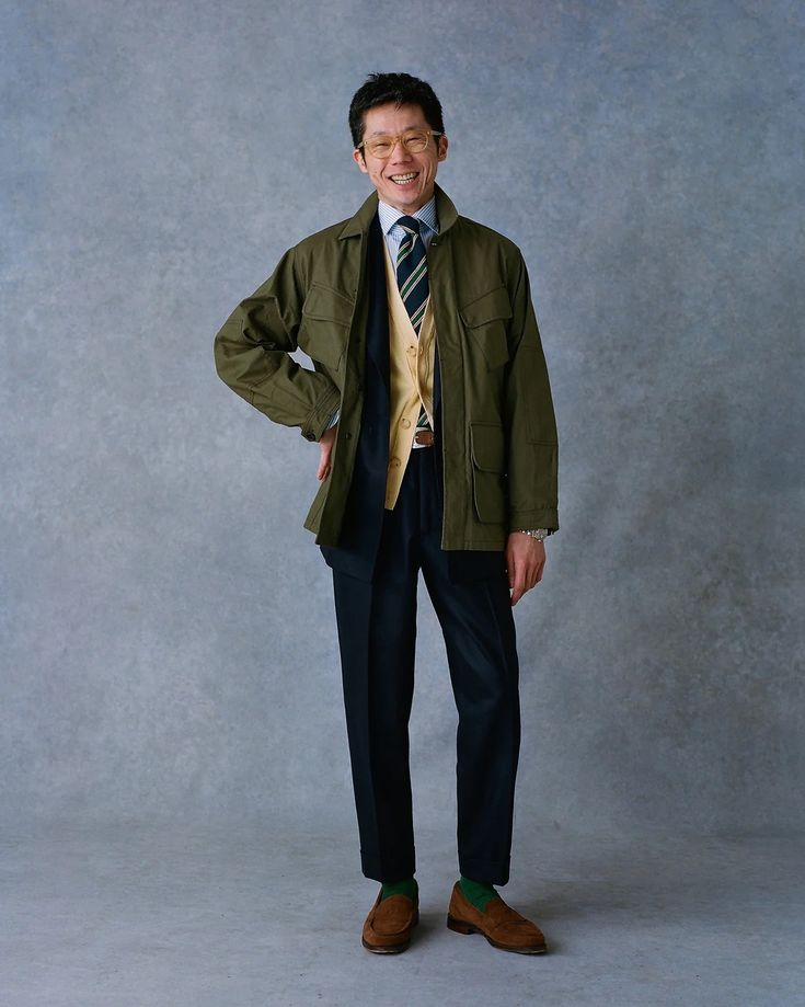 a fullbody studio shot of a smiling asian man wearing a stylish outfit. he's dressed in a dark olive green field jacket over a mustard yellow waistcoat and white shirt. he has a striped tie and dark navy blue trousers. his brown suede loafers and green socks complete the look.  the backdrop is a textured grey. he's standing with his hands on his hips, offering a friendly expression. the photo is welllit and the man appears confident and fashionable.