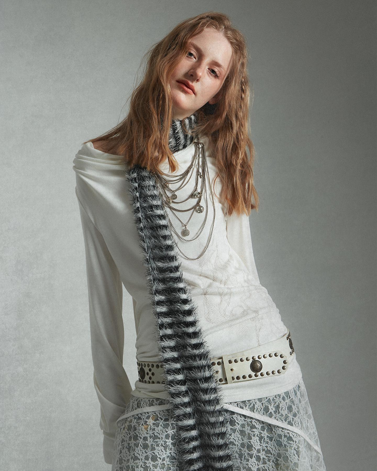 the image is a fashion studio portrait of a young woman. she is wearing a stylish offtheshoulder longsleeved white shirt layered over a white lace skirt.  a black and white fuzzy scarf is draped around her neck. she also wears a layered silver necklace and a wide white studded belt. her long blonde hair is styled in loose waves. the backdrop is a simple, neutral grey. the overall aesthetic is bohemian and chic.
