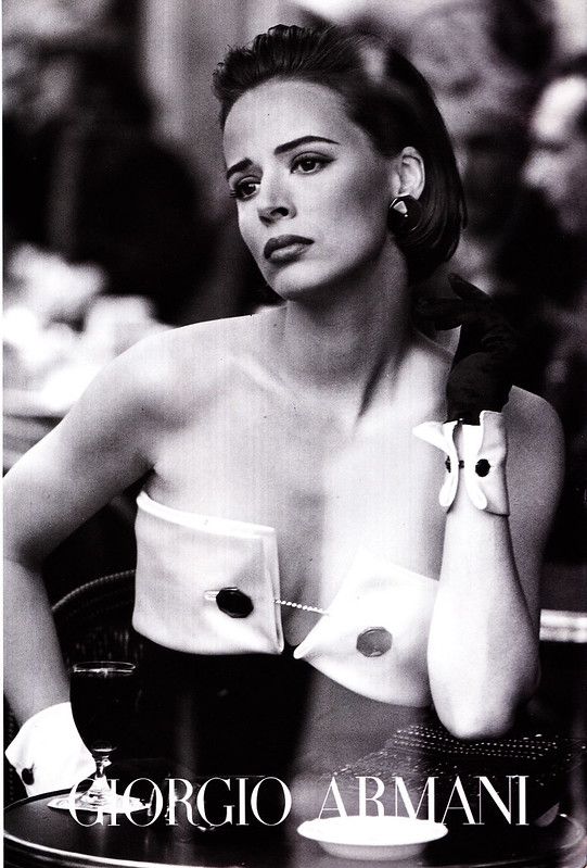 a black and white fashion advertisement for giorgio armani, featuring a woman in a strapless top secured by cufflinks, wearing gloves and earrings, sitting at a cafe table with a wine glass and a cup.