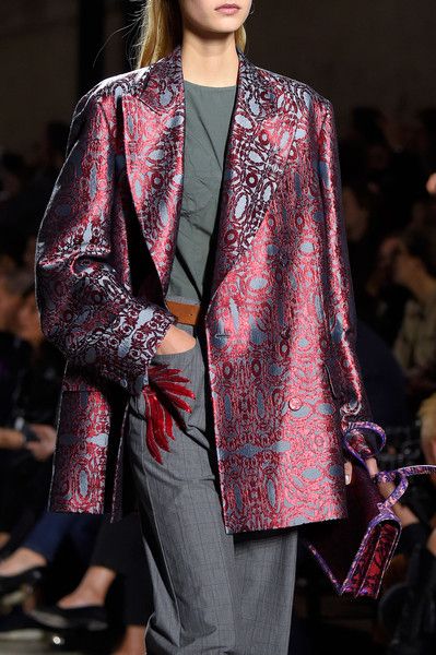 a closeup shot of a fashion model on a runway, showcasing a richly patterned red and blue brocade blazer. the model wears grey trousers and carries a purple handbag.  the background is blurred, but shows other people in the audience at a fashion show. the image emphasizes the texture and detail of the blazer.
