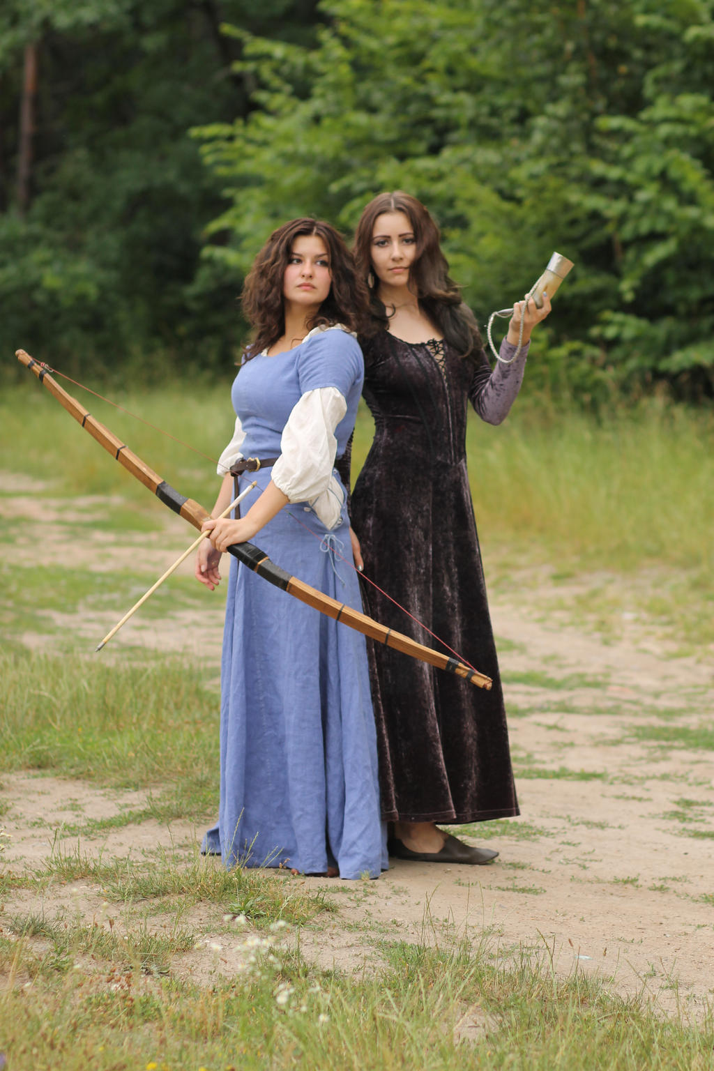 wo young women in medievalstyle costumes stand on a dirt path in a forest setting. one woman holds a longbow, while the other holds what appears to be a small horn or container.  they are dressed in long gowns; one in a blue dress and the other in a dark brown/purple velvet gown. the scene suggests a fantasy or historical reenactment setting.