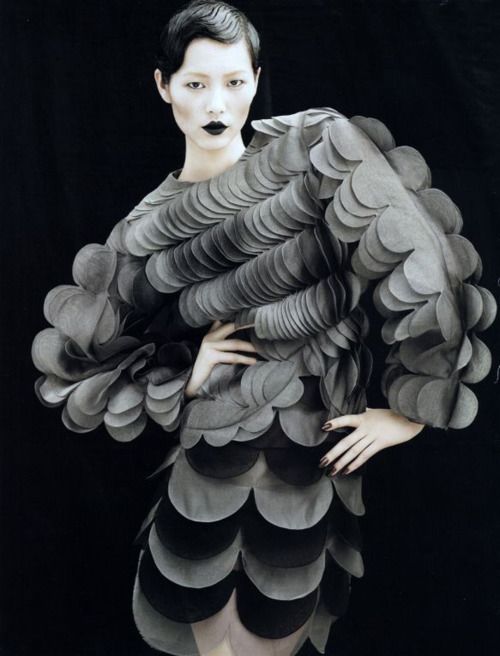 a woman with short black hair and pale makeup is wearing a gray,  avantgarde, couture, scallopshaped dress. the model poses and stares into the camera with her hands on her hips. the image has a dark, moody feel.