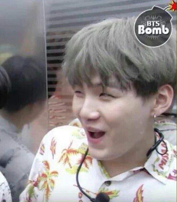 a closeup shot of suga from the kpop group bts, looking surprised with his mouth open. he's wearing a white hawaiian shirt and has gray hair. there's a microphone clipped to his shirt. the "bts bomb" logo is visible in the top right corner of the image.