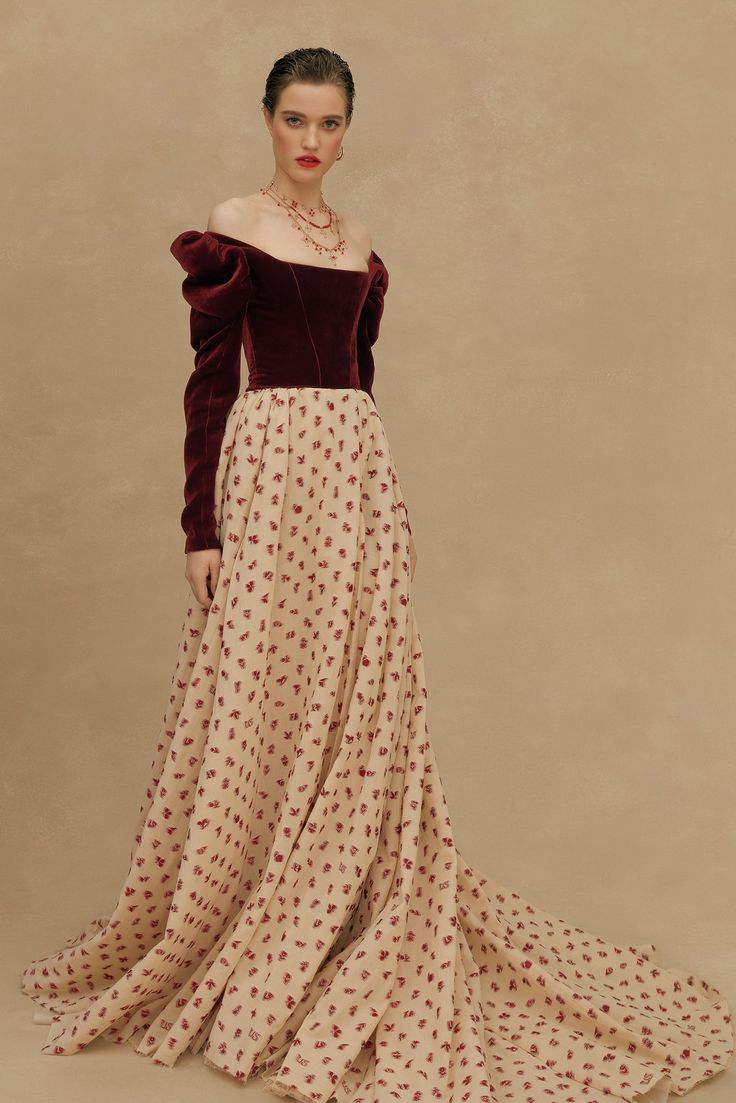 a fulllength portrait features a woman in an elegant dress against a neutral, beige background. the dress consists of a dark, burgundy velvet top with long, puffy sleeves that sit off the shoulder. the skirt is a light beige color and has a delicate red floral print throughout. the dress extends to the floor, with a slight train adding to its graceful appearance. the woman is wearing a necklace and has red lipstick on. her hair is neatly styled. the overall composition and color palette create a sophisticated and timeless aesthetic.