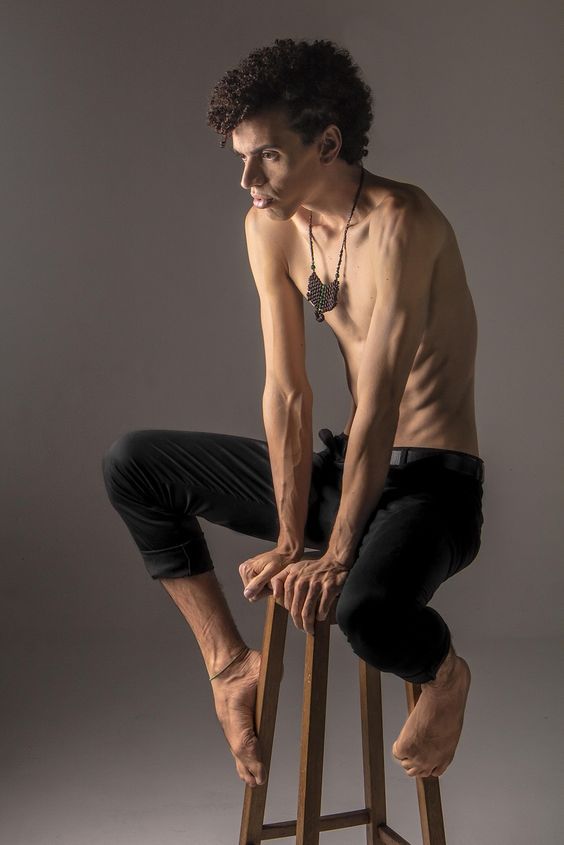 a young man in black pants is sitting on a wooden stool with his bare chest on display. he is looking away from the camera, his hair is curly and he is wearing a necklace.  the background is a light gray and the lighting is soft.  the man is skinny and muscular.
