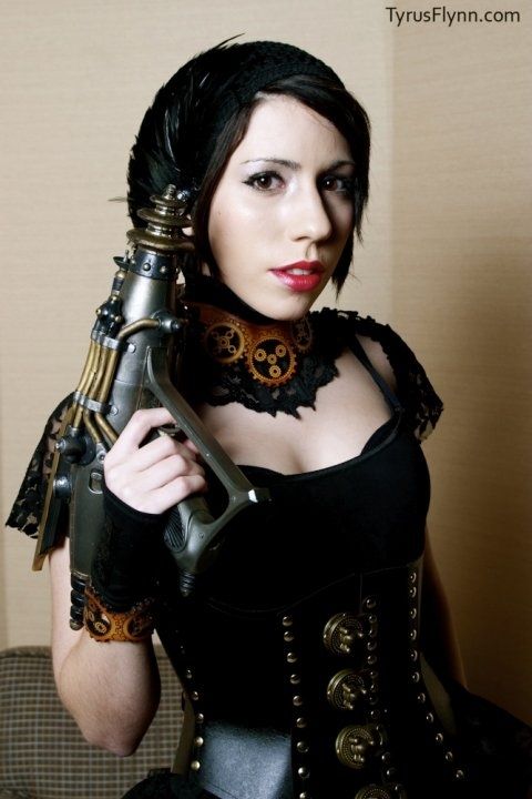 a woman in a steampunk costume holds a steampunk gun in her hand. she is looking directly at the camera with a serious expression. the gun is a complex design of metal and gears. she is wearing a black corset and a choker necklace with gears.