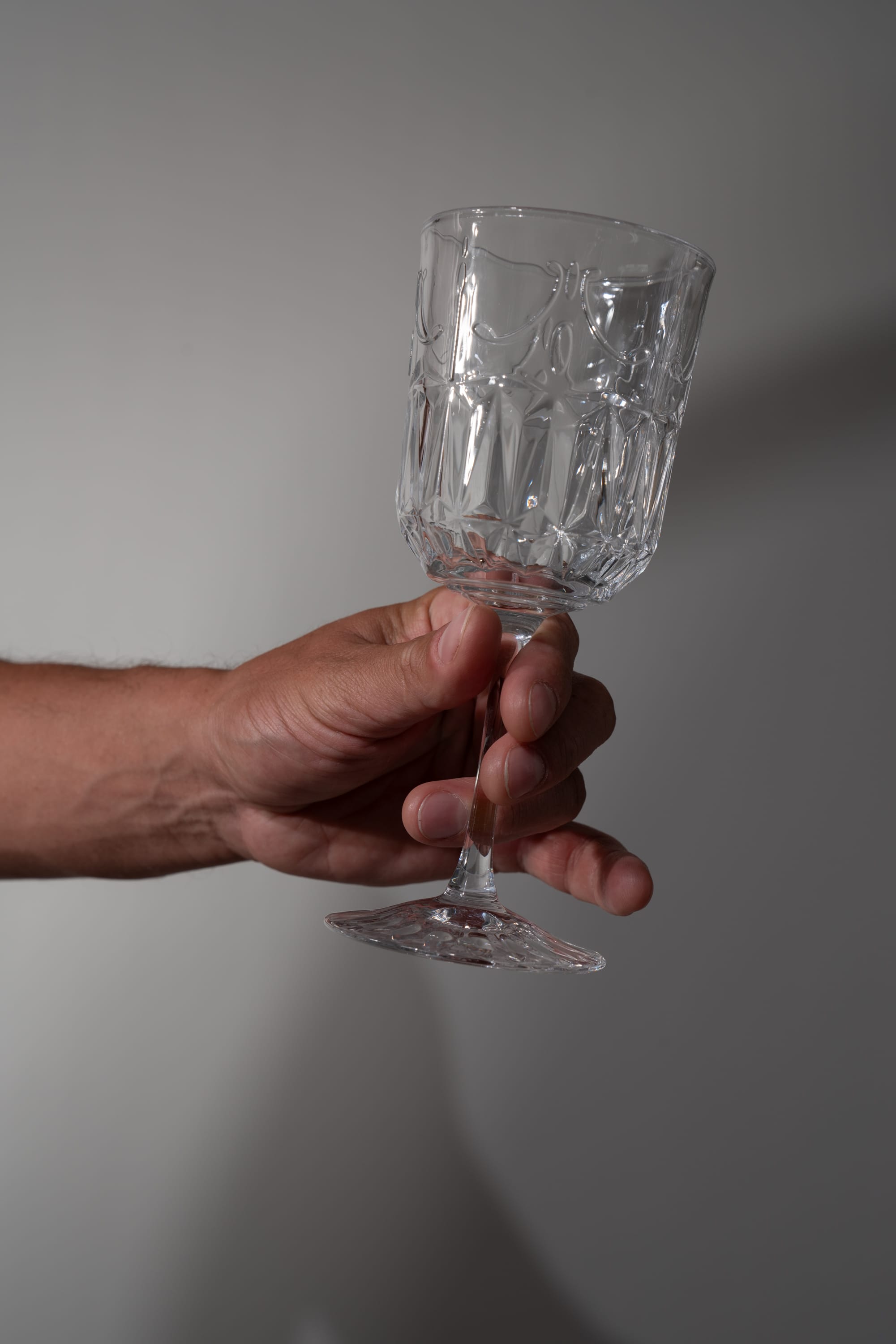 a man holds an empty crystal wine glass against a light gray background. there are subtle shadows cast by the hand and glass on the wall, hinting at a light source from the left.