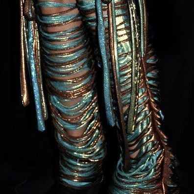 the image shows a closeup of a person's legs adorned in a very elaborate and artistic legwear. the legwear consists of spiraling golden cords overlaid on what appears to be blue stockings. the cords and ropes are arranged to create a somewhat gladiatorlike aesthetic, giving the legwear a costumelike quality. the metallic and iridescent nature of the materials suggests a theatrical or fashionforward setting.